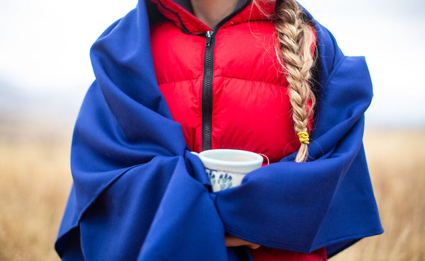 A person wearing a bright red down vest, standing outdoors on an overcast winter day; their hair is in a long braid and they are holding a mug of hot tea. The Shepherd's Wool in Neela Blue is wrapped around their shoulders and hands for warmth.